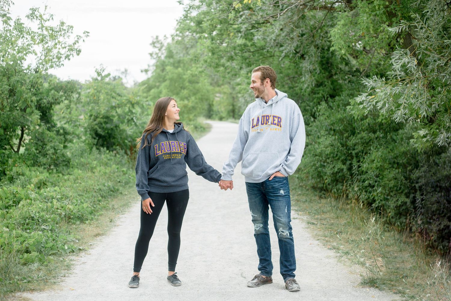 Couple holding hands and smiling at each other wearing Laurier hoodies on a tree-lined gravel path.
