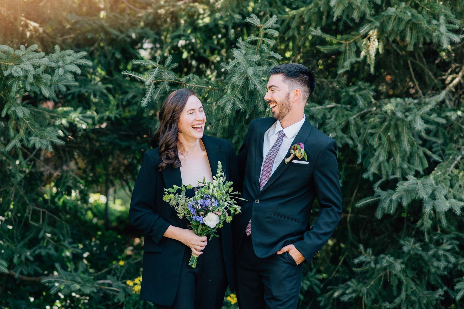 Couple in black attire smiles at each other holding bouquets before lush green evergreen trees outdoors.