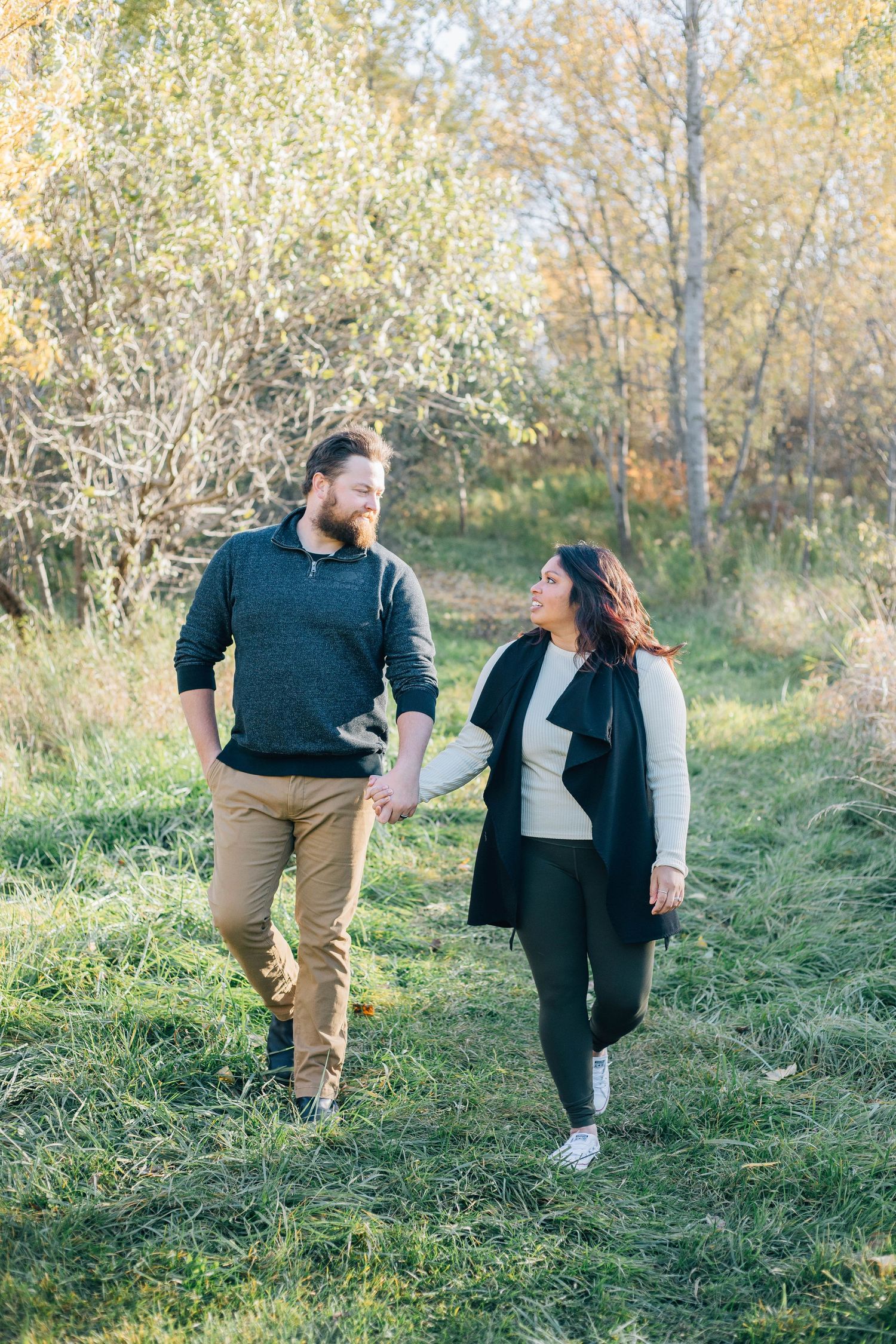 Couple holding hands and smiling at each other while walking through a sunny autumn meadow.