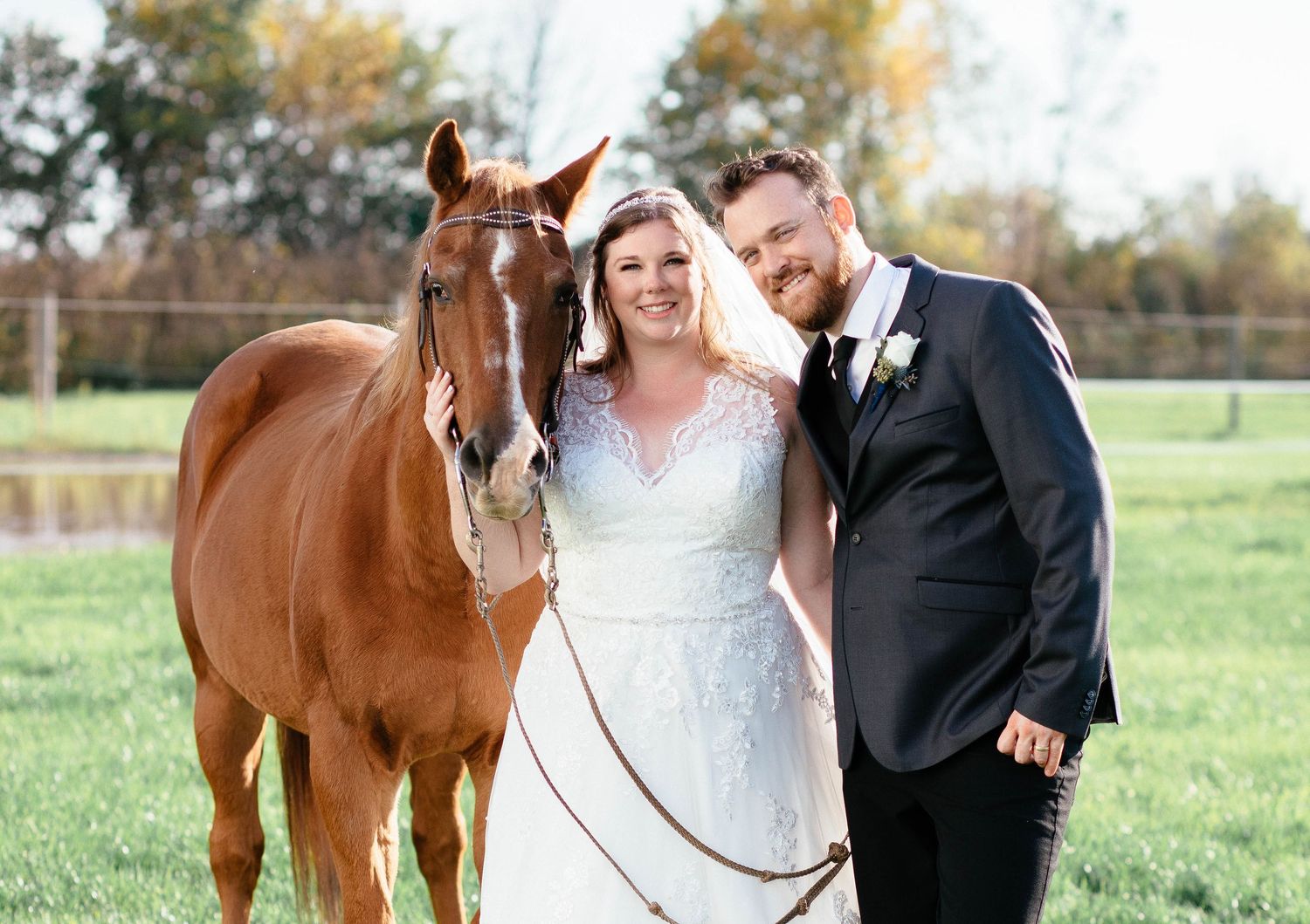 Smiling bride and groom pose with chestnut horse in green pasture on their autumn wedding day.