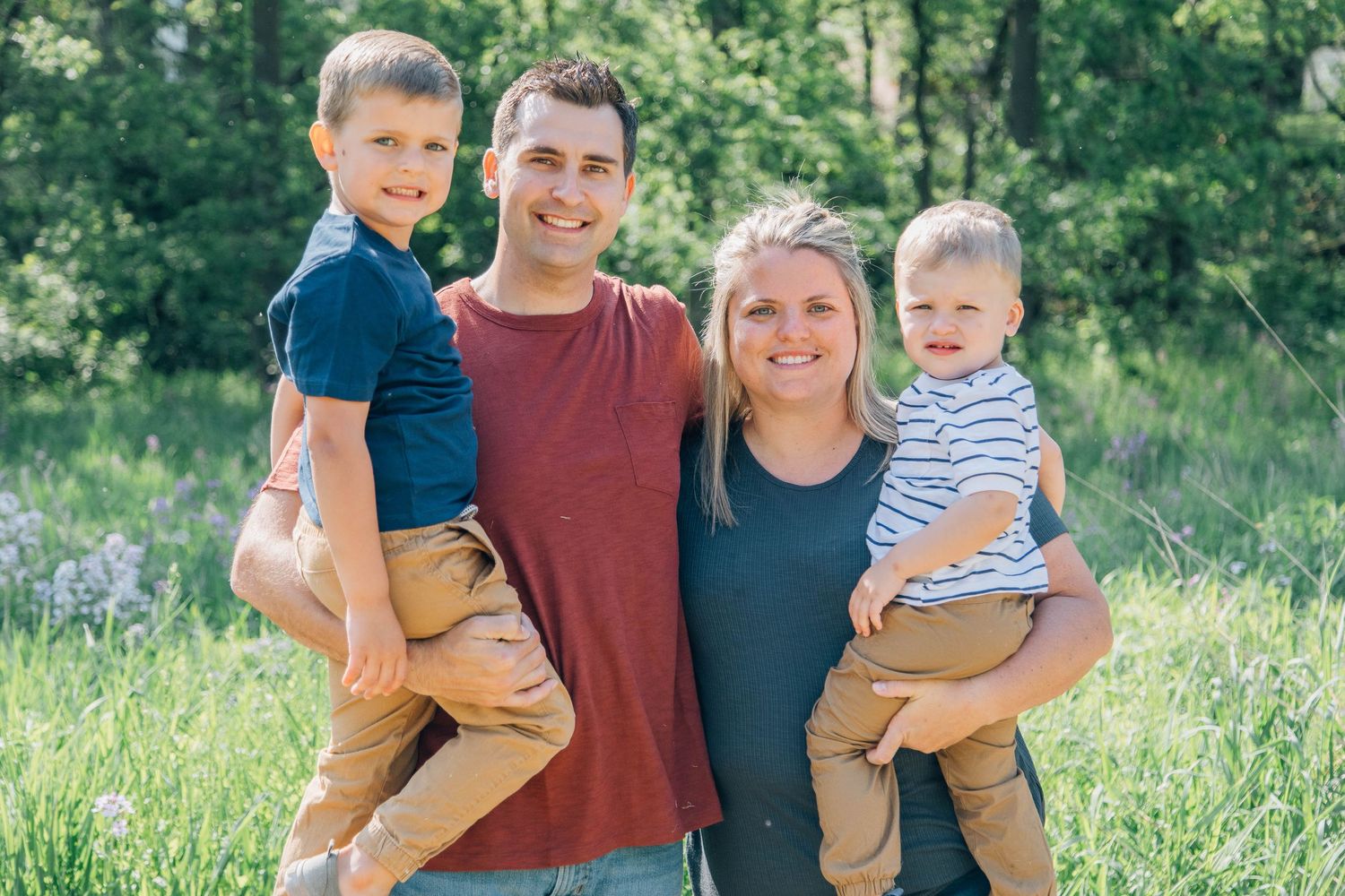 Happy family of four with parents each holding a young child in a green meadow.