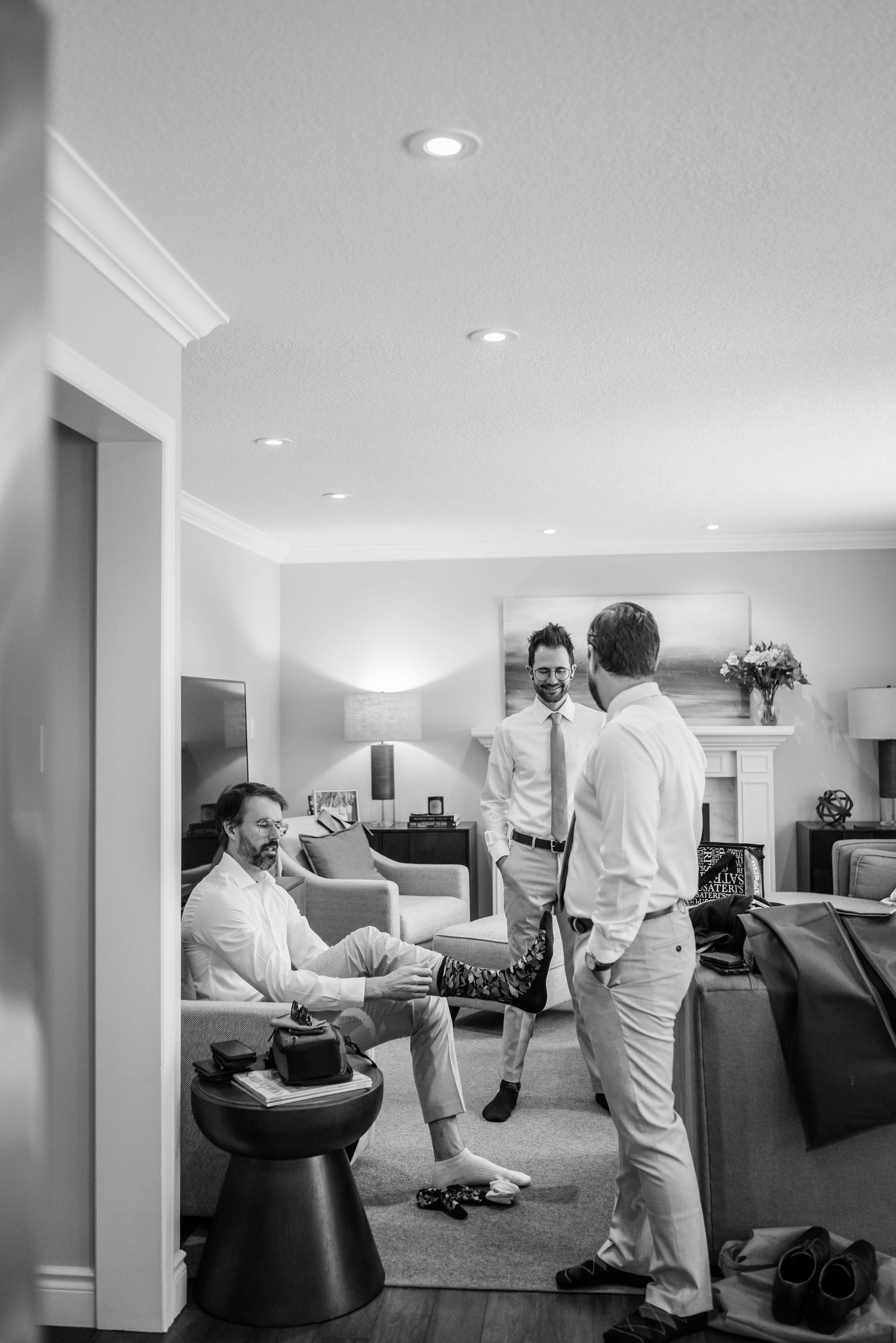 Black and white photo of groomsmen chatting and getting dressed in a bright living room.
