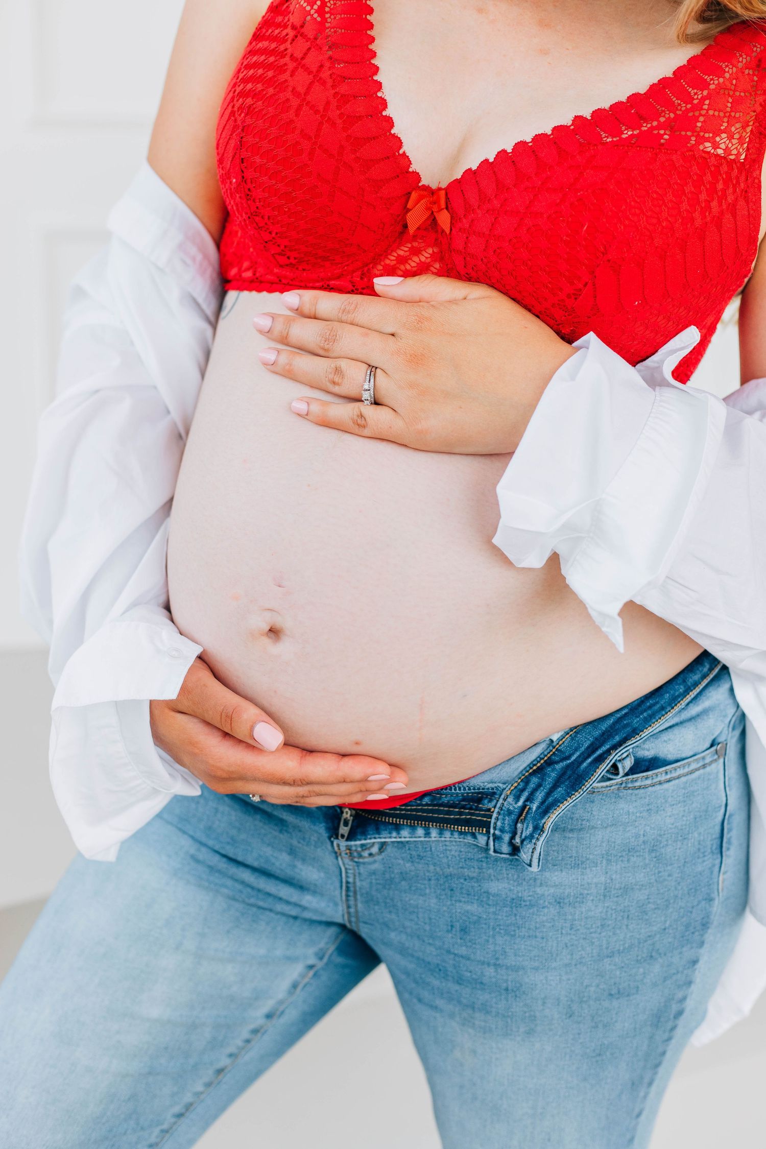 Close-up of pregnant woman in red bralette and jeans gently holding her bare baby bump.