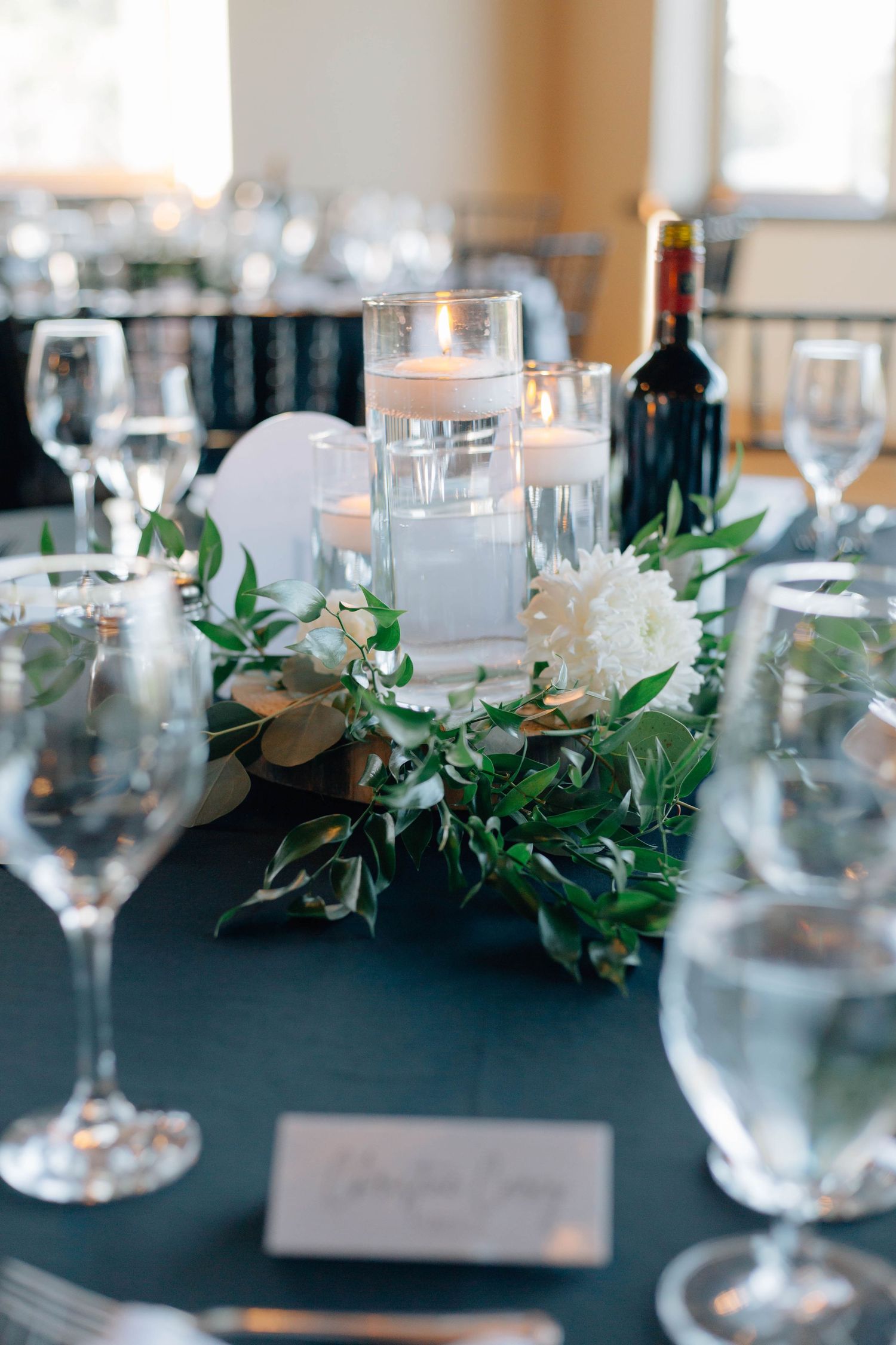 Elegant wedding table centerpiece with floating candles, white flowers, and greenery on dark teal tablecloth.