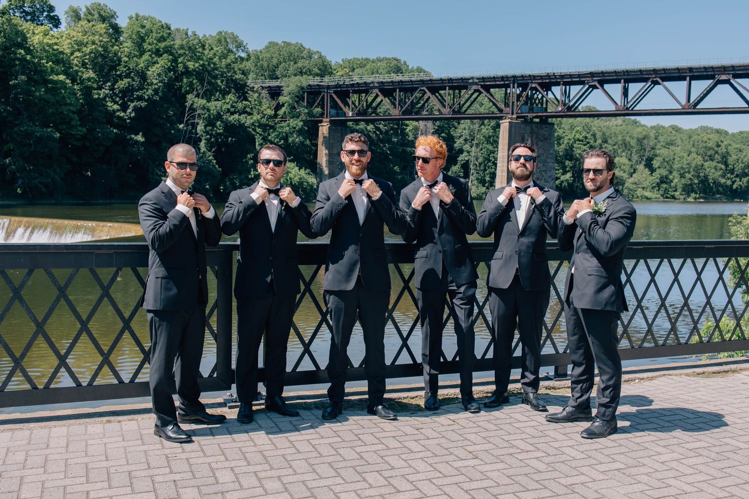 Six groomsmen in black suits posing playfully by a metal railing with a river and bridge in the background.