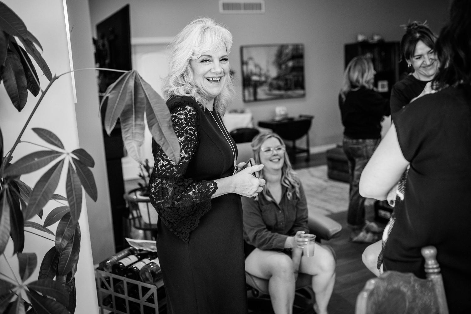 Elegant blonde woman in black lace dress laughing with bridesmaids during wedding preparations.