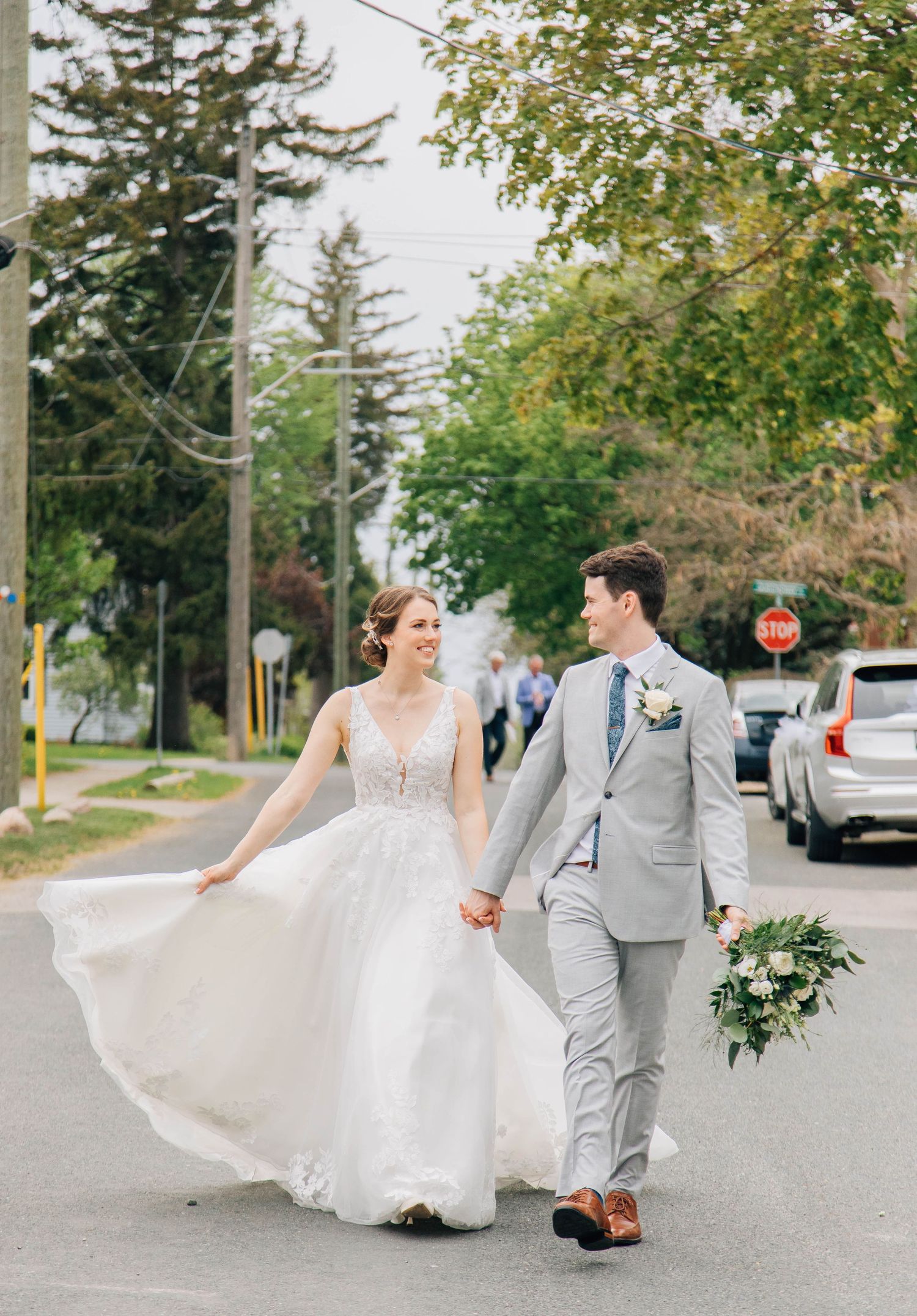 Bride and groom walk hand-in-hand on a sunny street in St Jacob's Ontario, groom in gray suit holding bouquet.