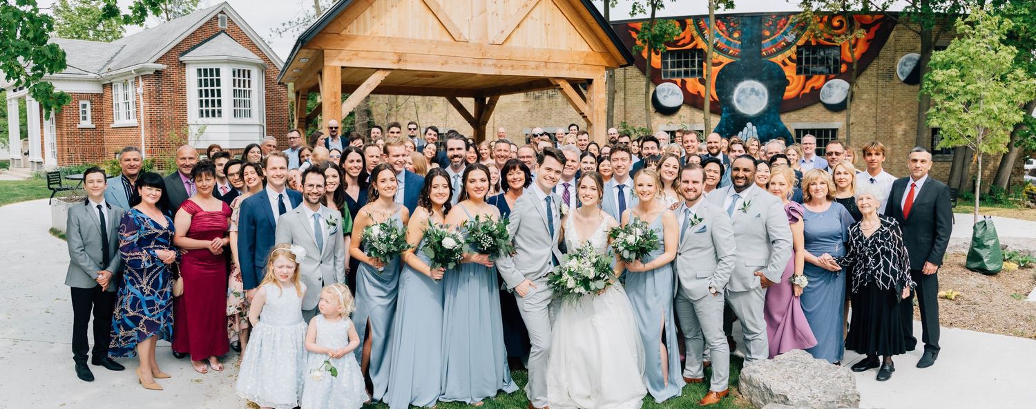 Large wedding party and guests posing together outdoors in front of rustic venue.