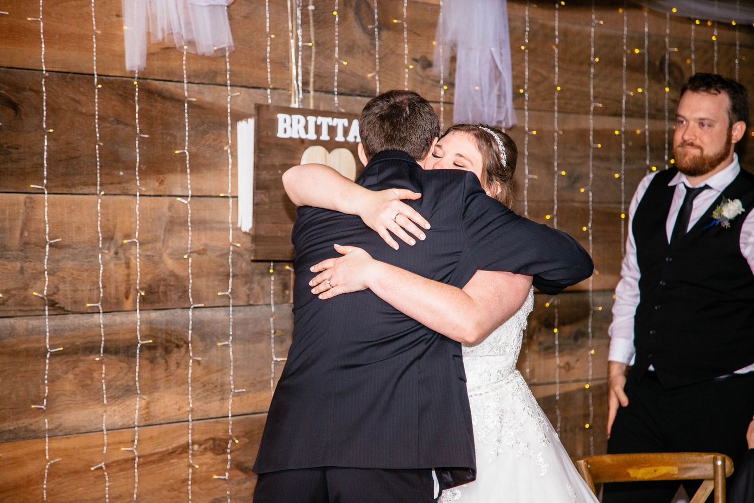 Bride sharing emotional hug with guest at rustic barn wedding reception near name sign.