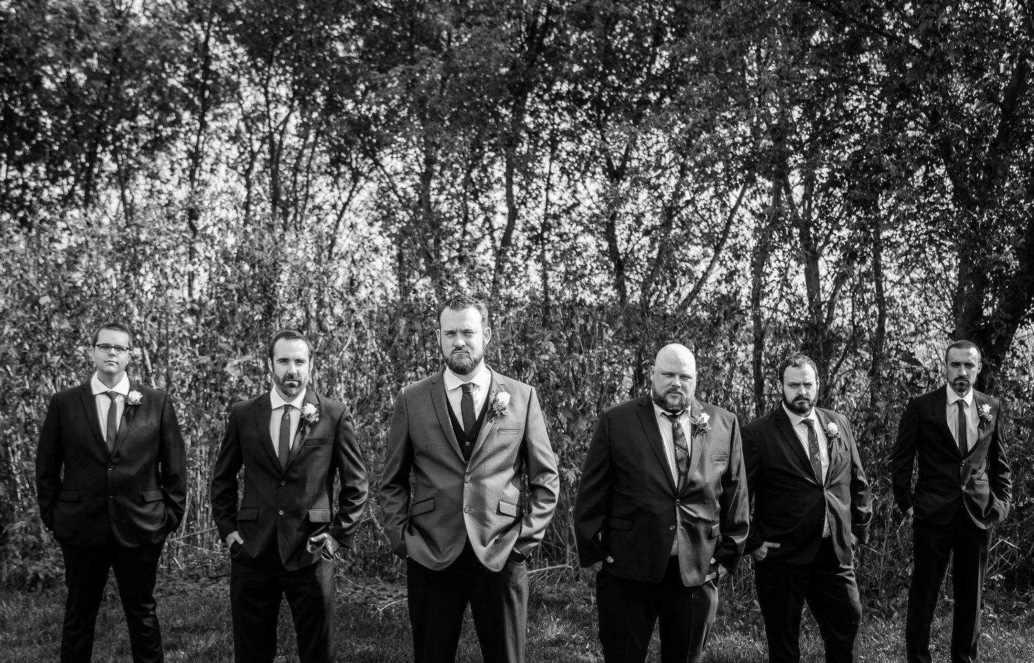 Black and white photo of groom and groomsmen in suits posing seriously among trees at wedding.