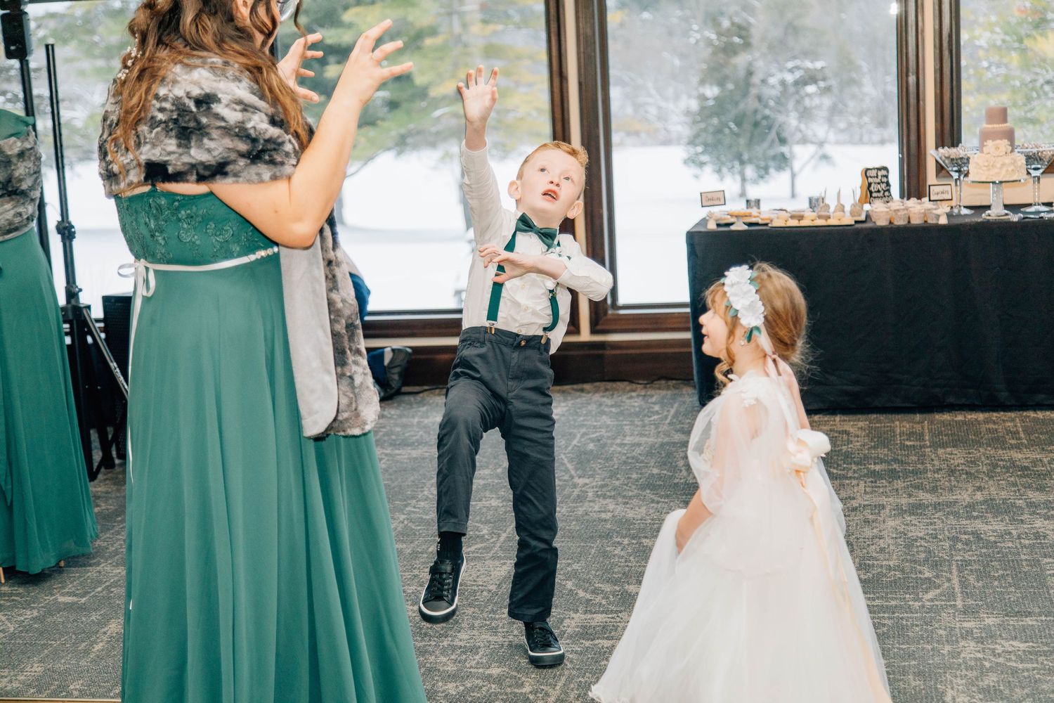 Ring bearer in suspenders and flower girl in white cape dress posing together in snowy winter landscape.