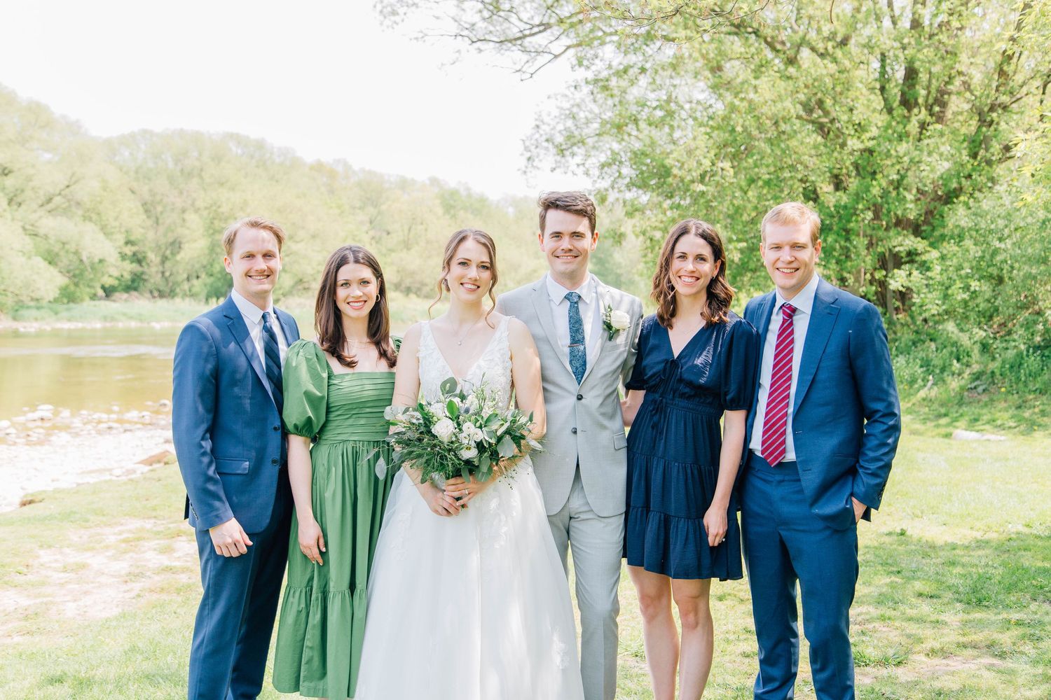 Wedding party of six smiles together outdoors by a river on a sunny spring day.