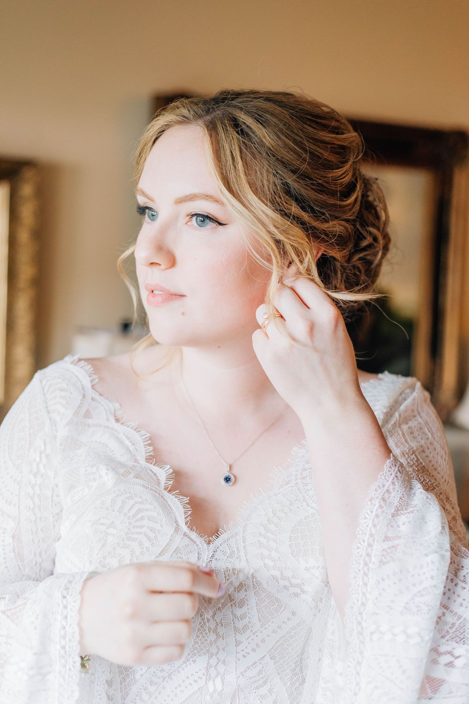 A beautiful bride with an elegant updo adjusts her earring while wearing a lace dress before her wedding ceremony.