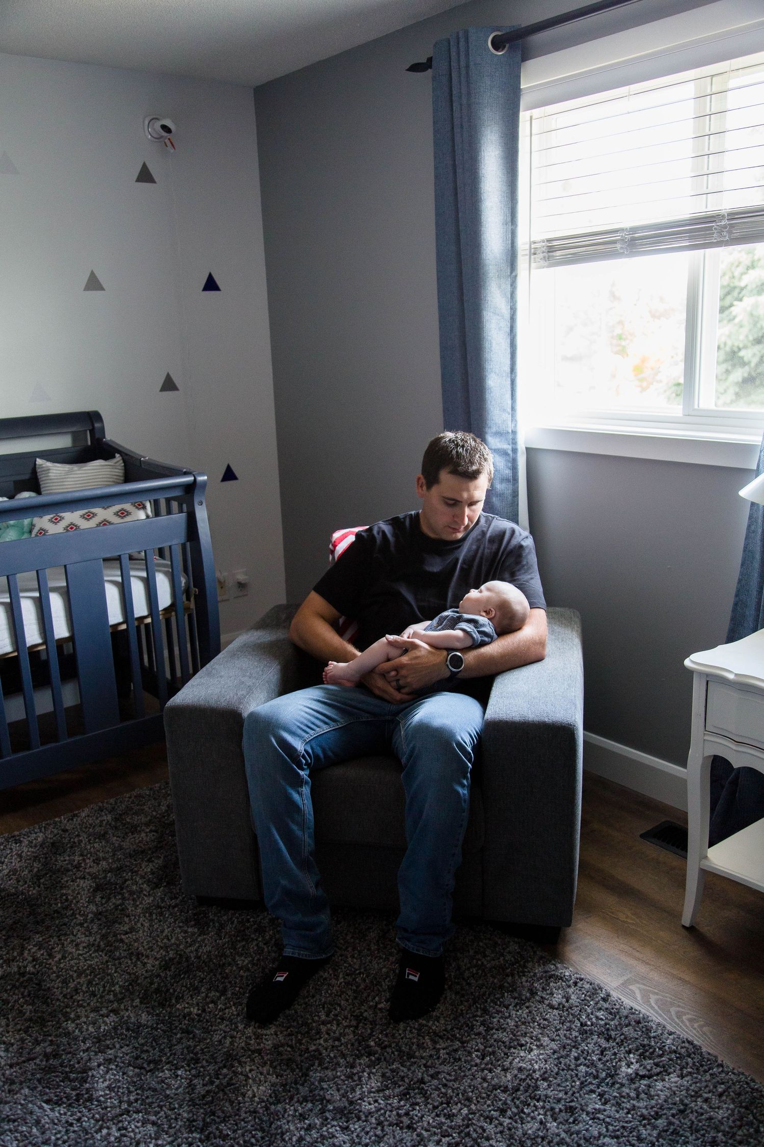 Father sits in nursery chair cradling his newborn baby by the window.