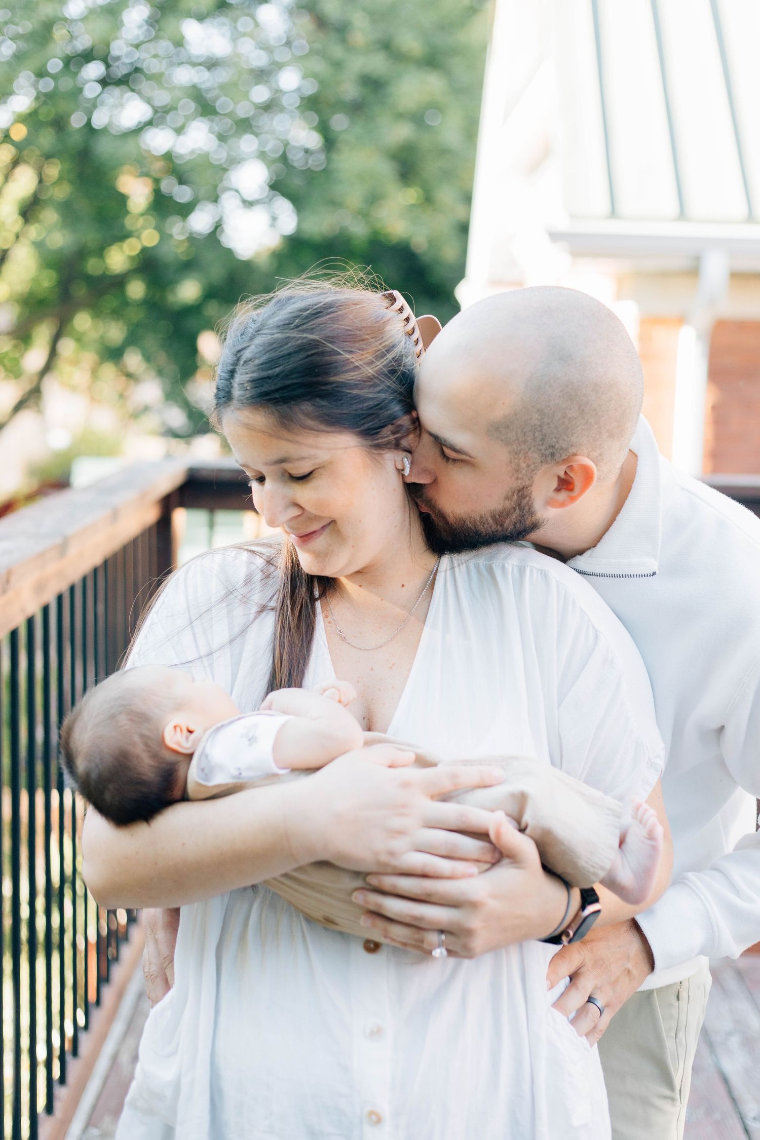Tender moment as man kisses woman holding newborn baby on sunlit outdoor deck.