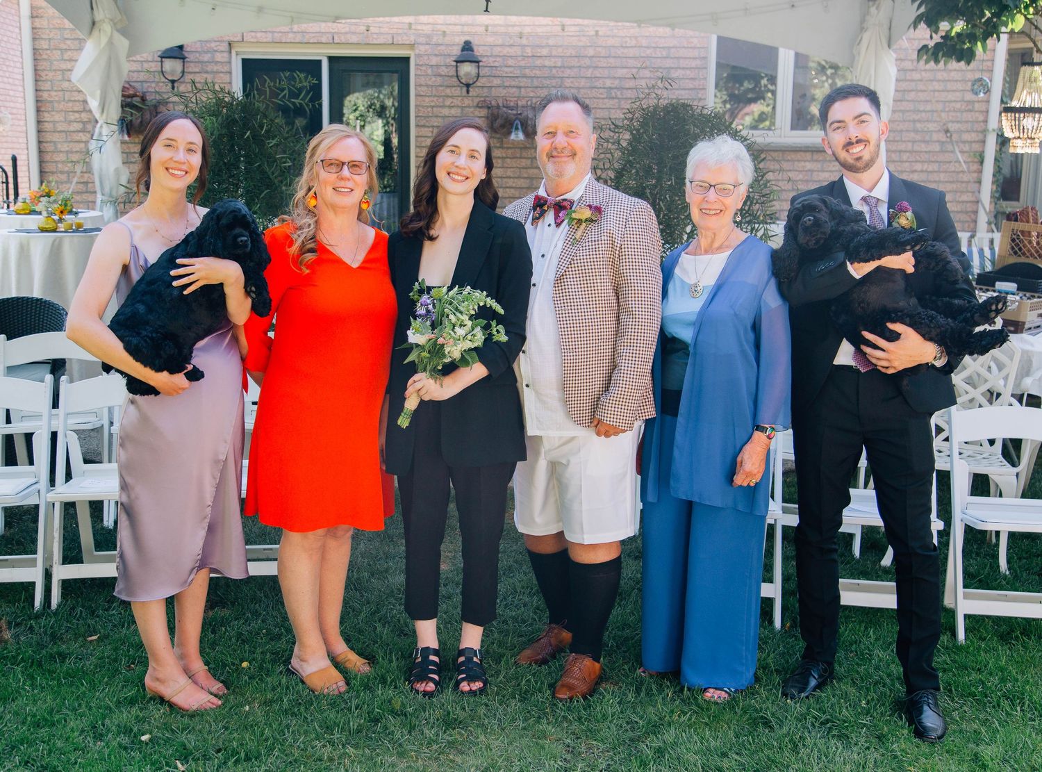 Wedding party of seven people posing together outdoors, including two brides and family members holding small black dogs.