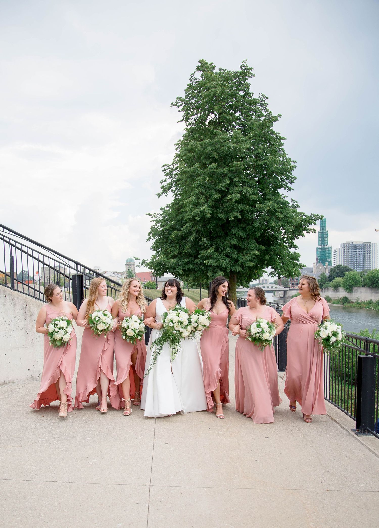 A bride in white walks with six bridesmaids in dusty rose gowns carrying white bouquets outdoors.