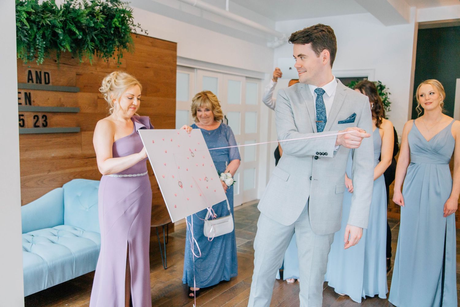 Groom reacts joyfully to wedding poster shown by smiling bridesmaid while bridesmaids in blue dresses look on.