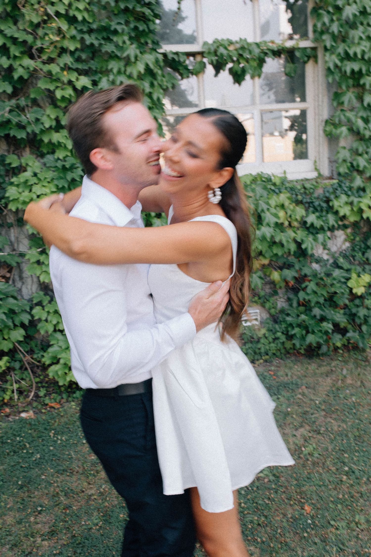 Happy couple embraces and laughs together in front of lush ivy-covered wall outdoors.