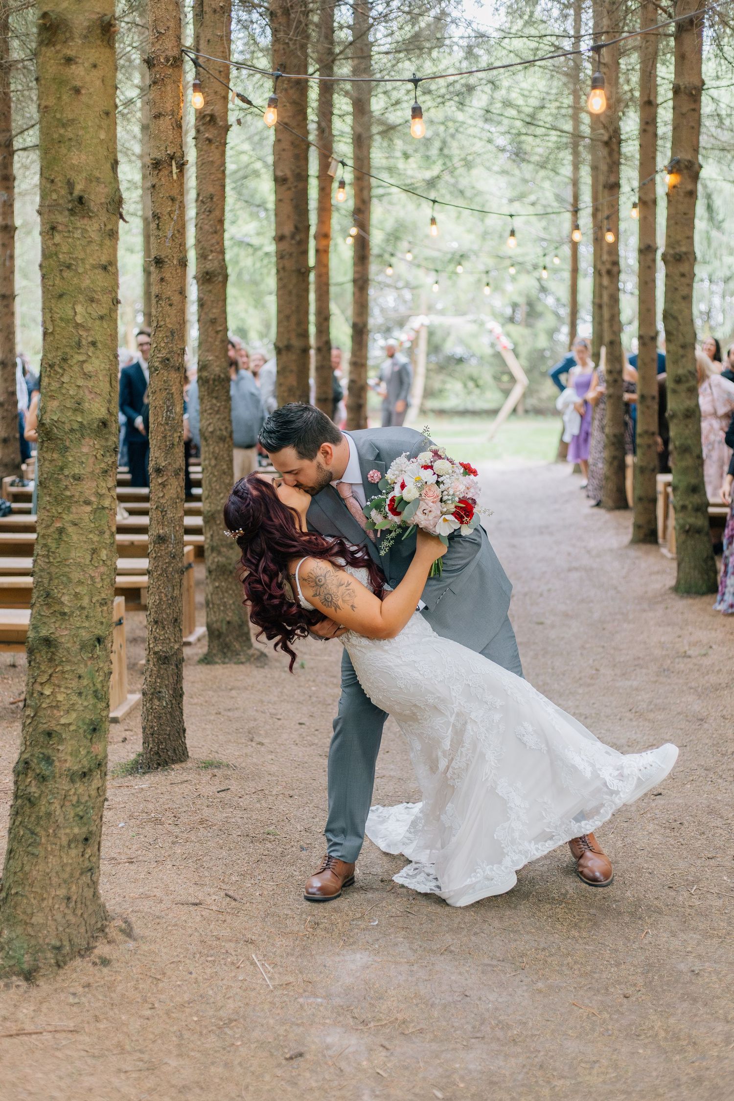 Groom dipping bride for a kiss along a pine tree aisle with string lights at Crescent Hill Acres.