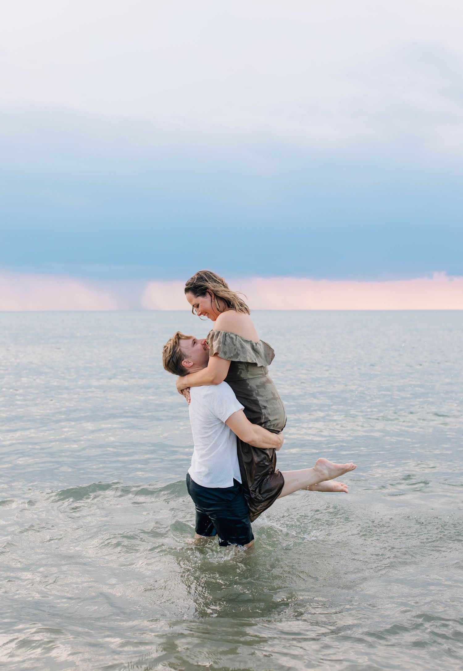Couple embracing in ocean waves at sunset, man lifting woman as they laugh together at Pinery Provincial Park.