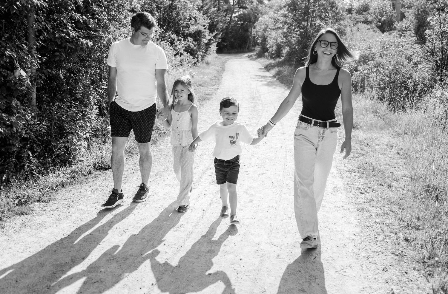 Black and white photo of a family of four walking hand-in-hand down a sunny dirt path surrounded by trees.
