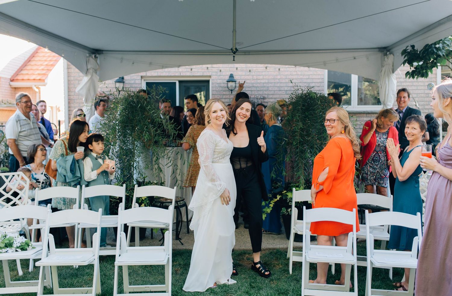 Bride in white lace gown walks down aisle with guests cheering at outdoor wedding reception.