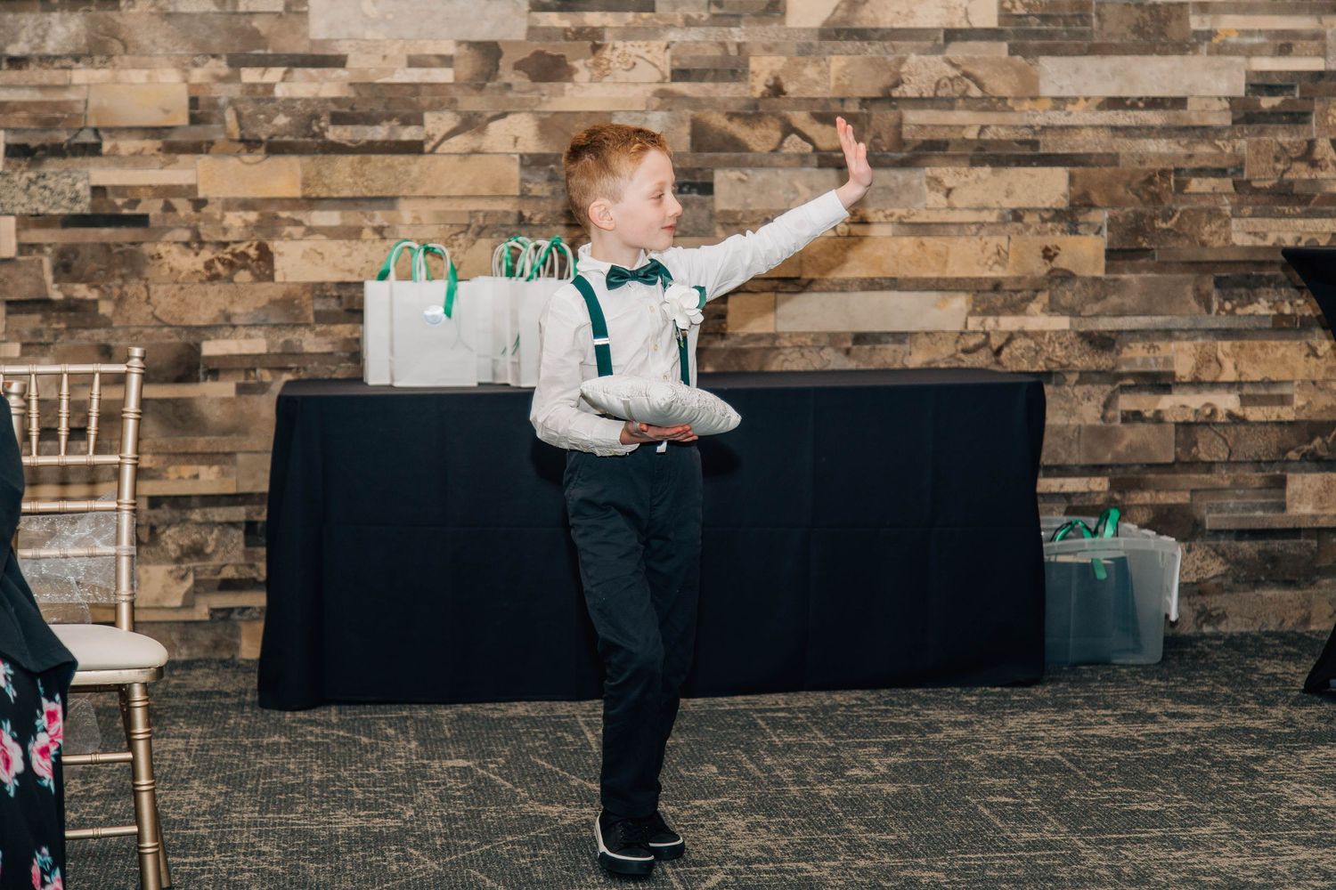 Young ring bearer in suspenders and bow tie dances energetically during wedding ceremony in decorated venue.