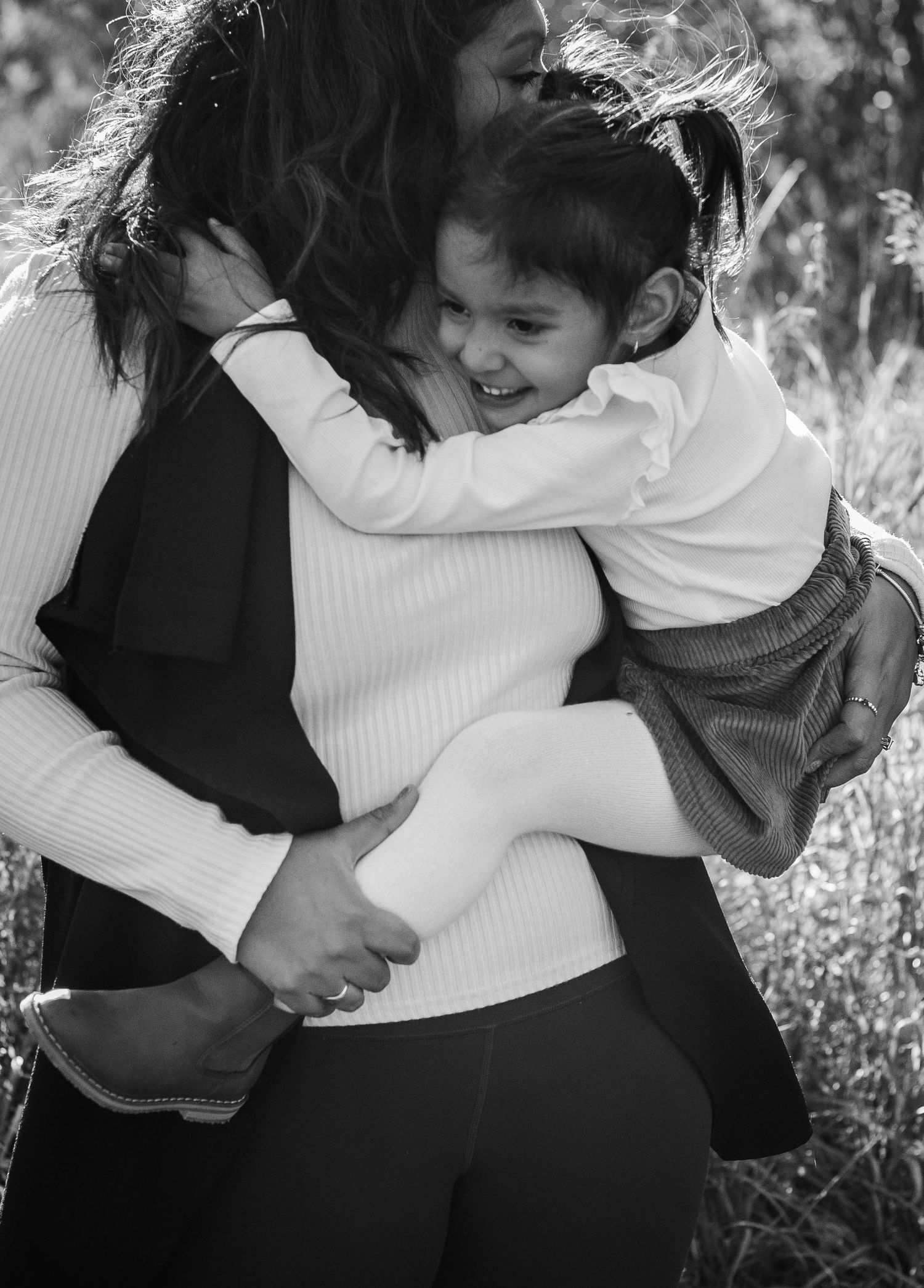 Black and white photo of young girl laughing while being hugged and carried by her mother outdoors.