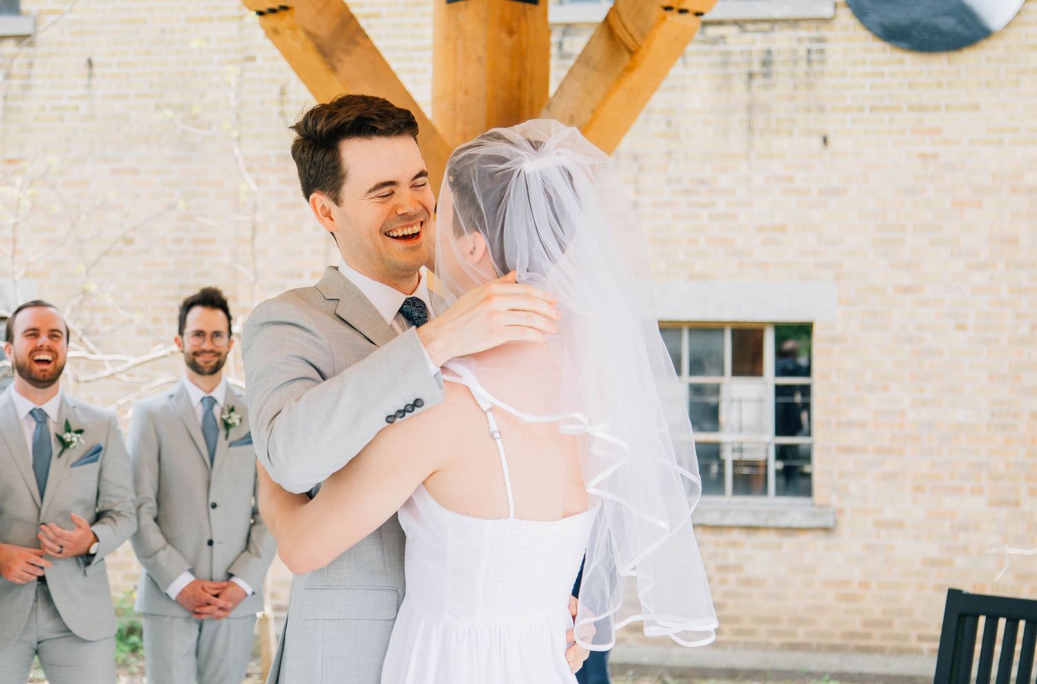 Joyful groom in gray suit embraces bride in white dress during outdoor wedding ceremony.