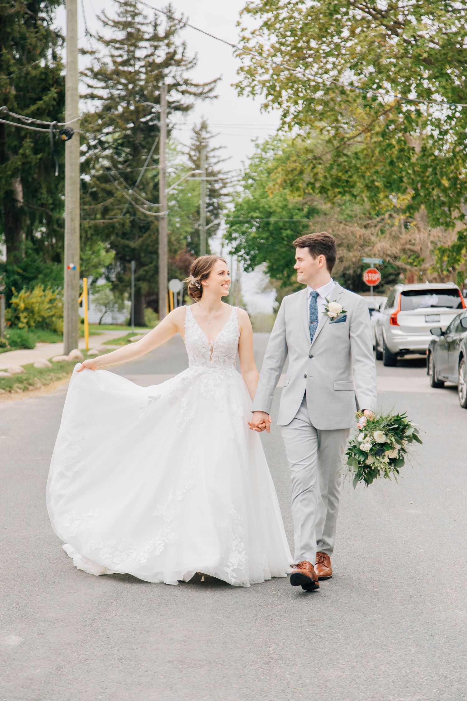 Bride and groom walking hand in hand down a tree-lined street, brides gown flowing in the breeze.
