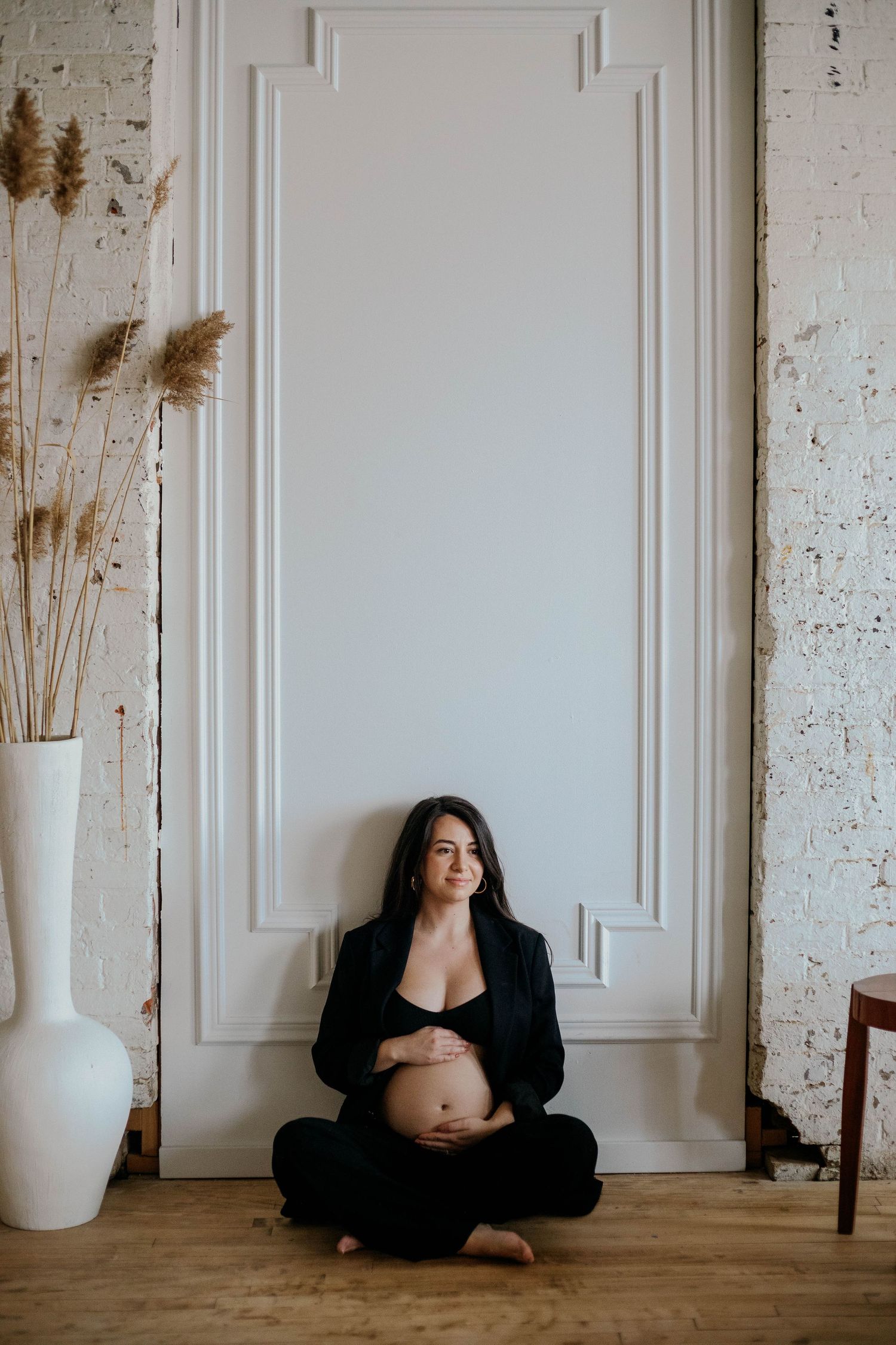 Pregnant woman in black outfit sitting cross-legged on floor, holding belly near decorative dried pampas grass.