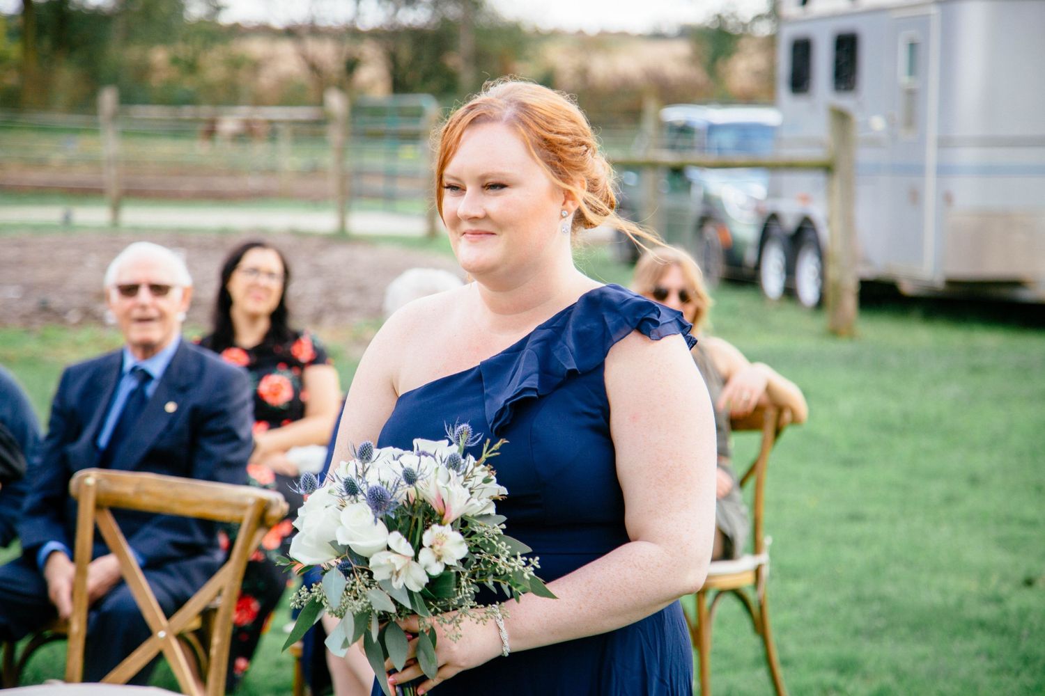 Bridesmaid in a navy one-shoulder gown holds a white floral bouquet while smiling at an outdoor farm wedding ceremony.