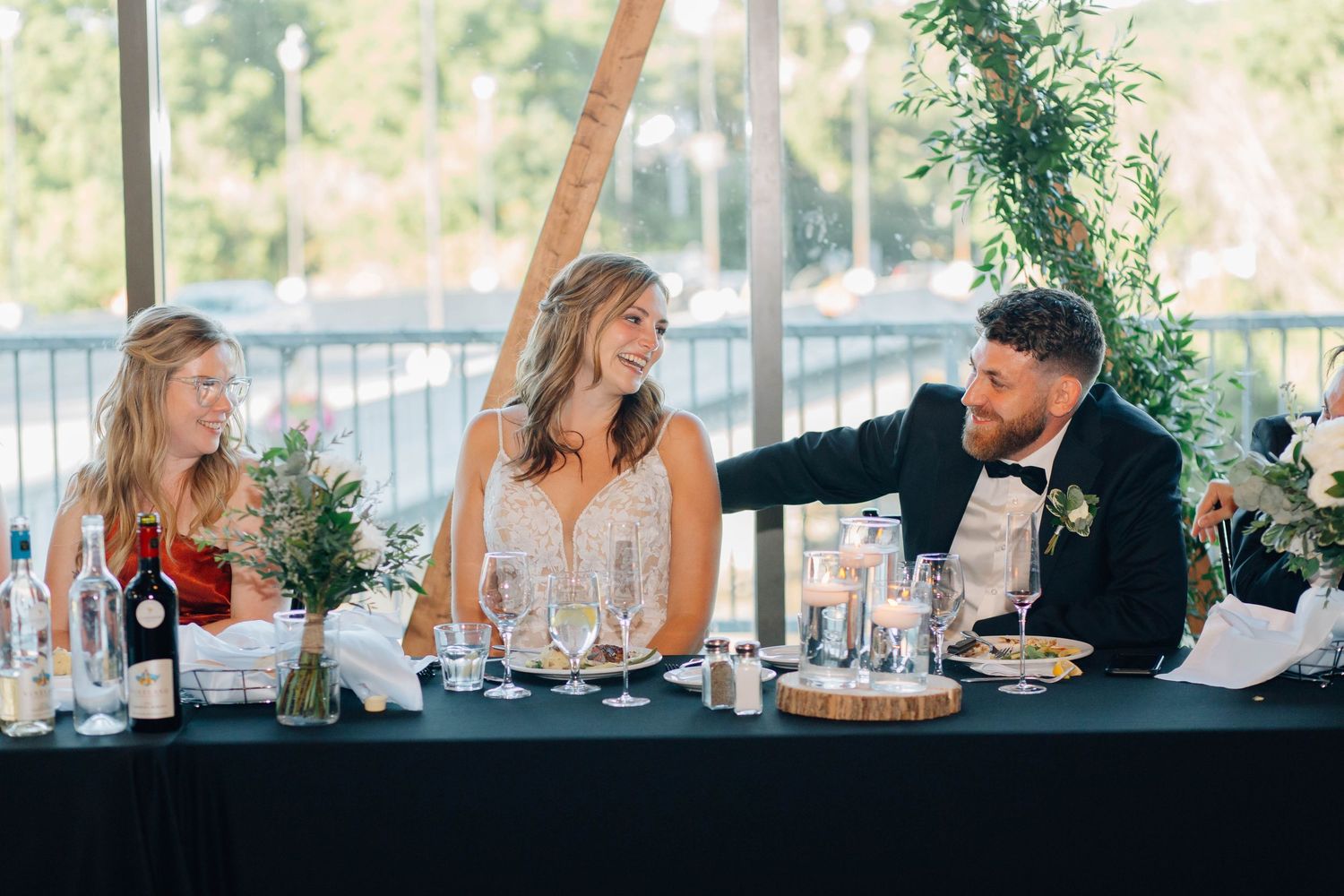 Bride laughs joyfully with guests at wedding reception head table decorated with candles and greenery.