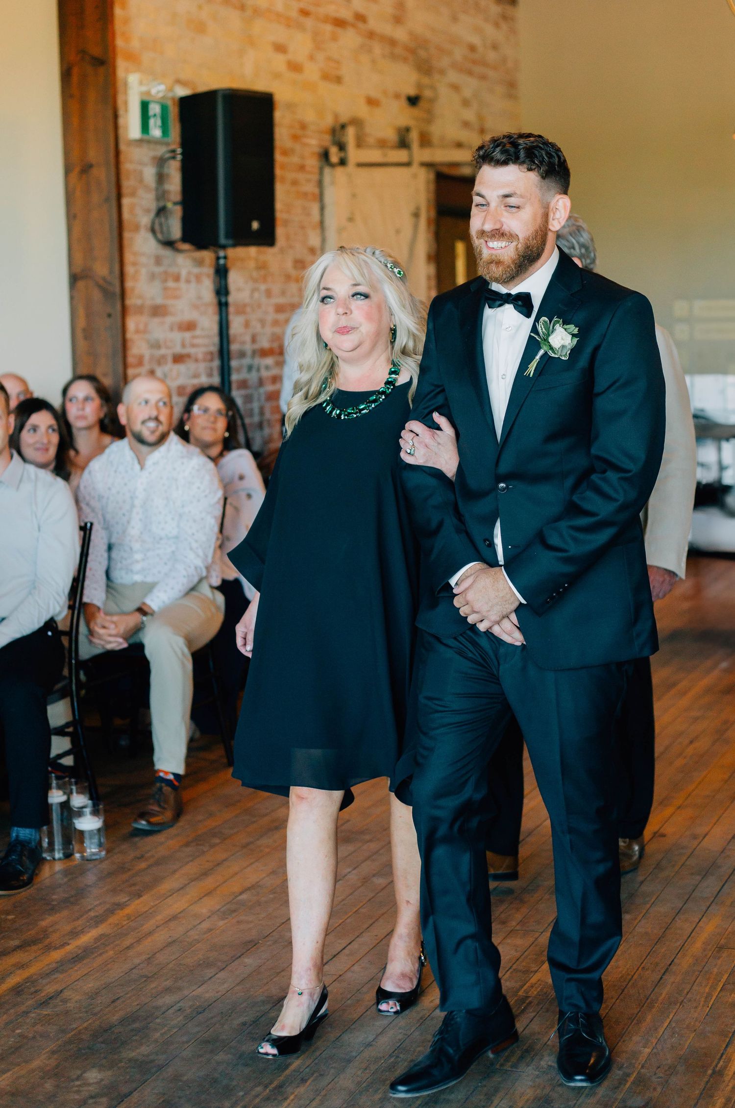 Groom in black tuxedo escorts smiling woman in black dress down wedding ceremony aisle.