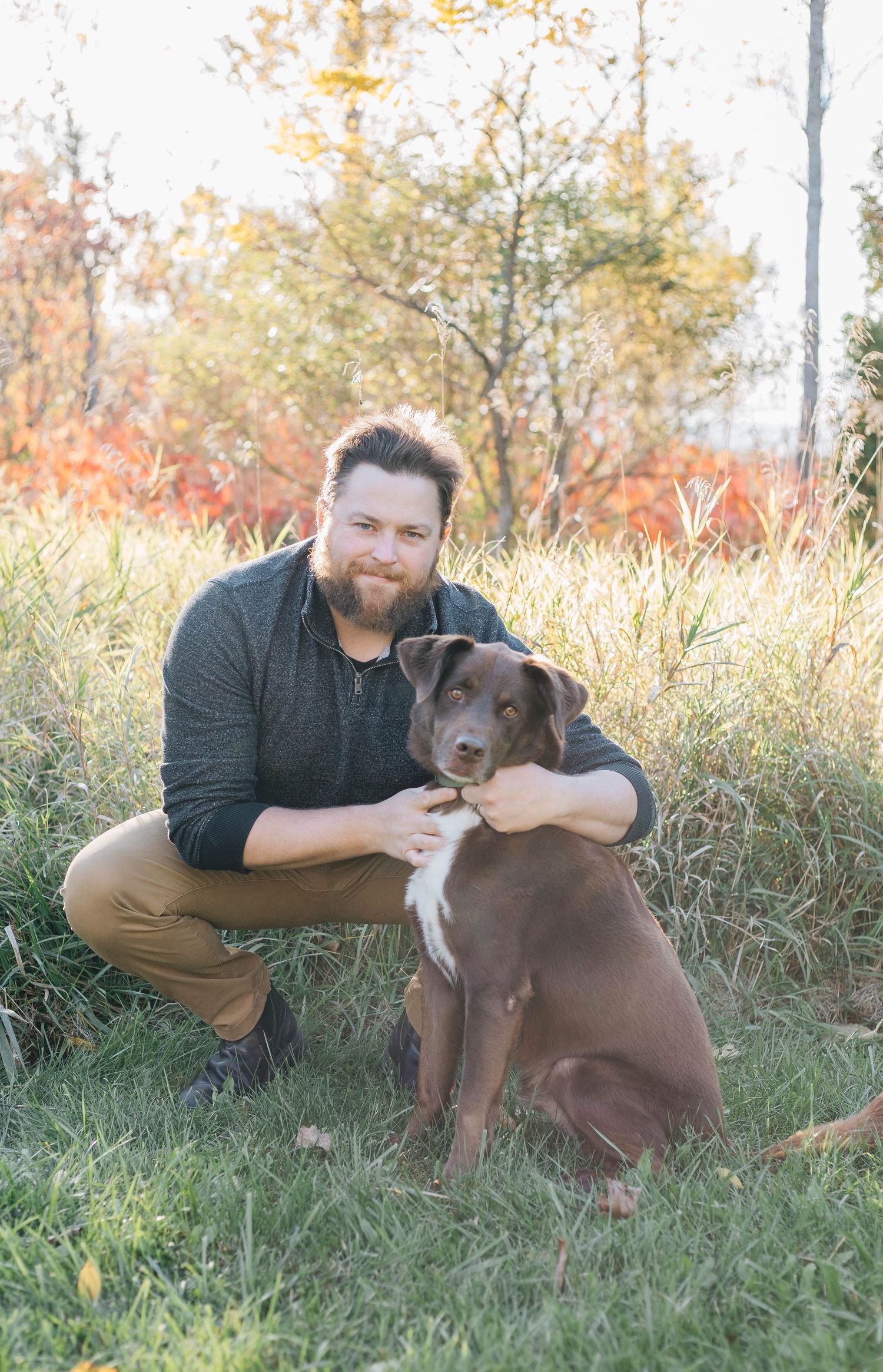 Bearded man crouching in autumn grass hugging a chocolate Labrador mix dog outdoors.