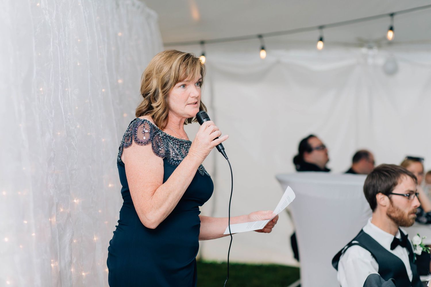 Elegant woman in navy lace dress delivers heartfelt speech with microphone at wedding reception.