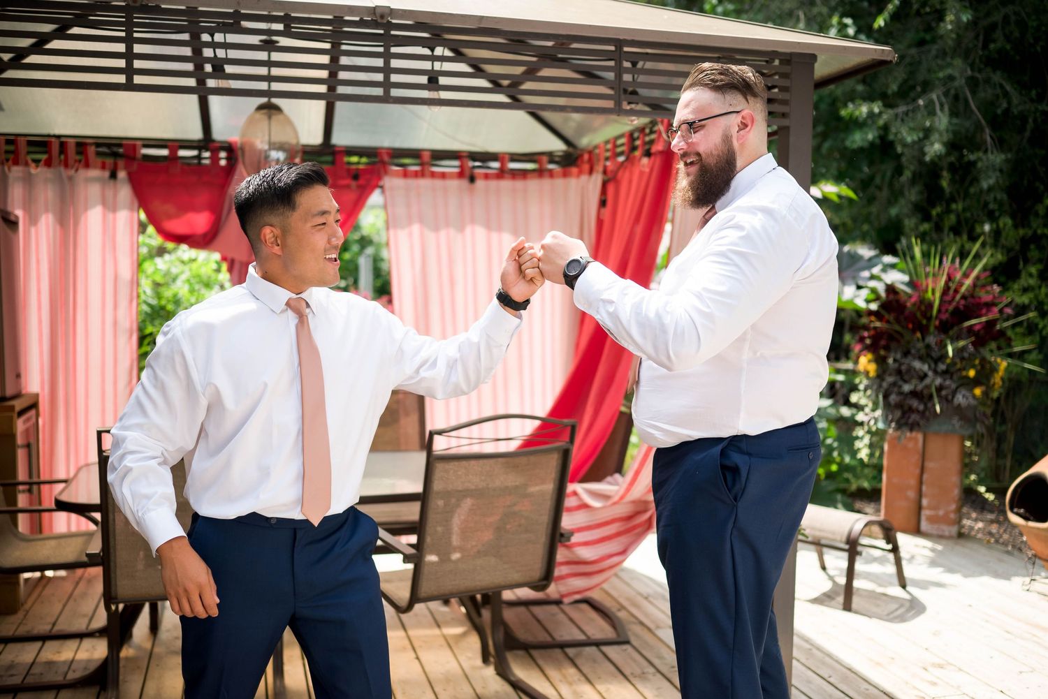 Two groomsmen in white shirts and navy pants celebrate with a fist bump on an outdoor deck.