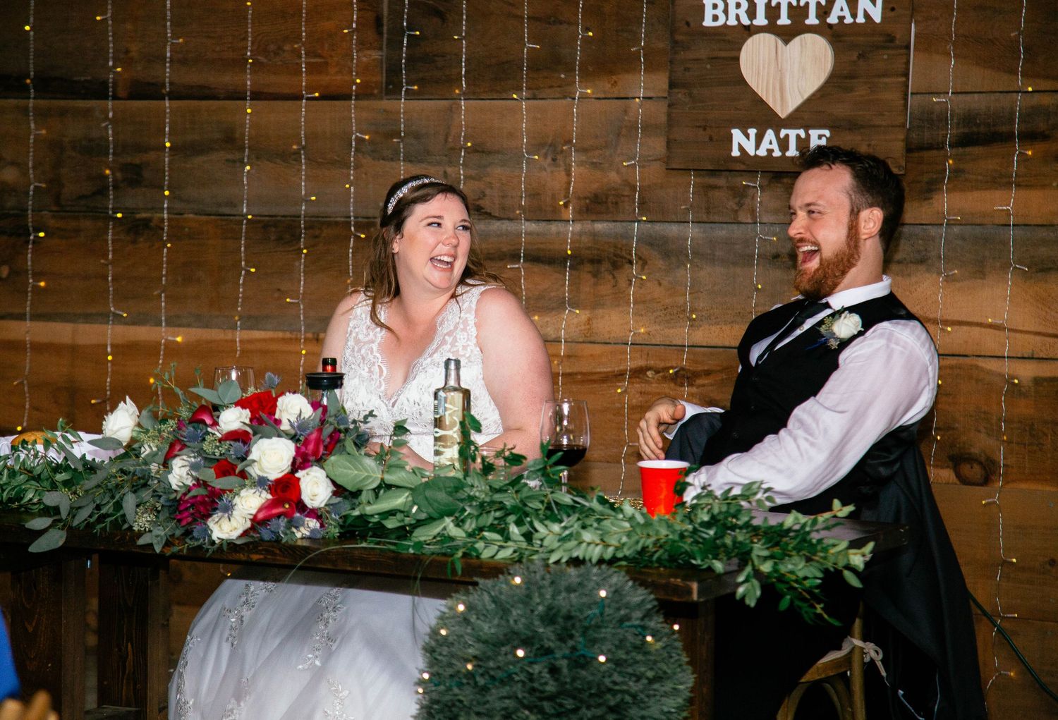 Bride and groom laughing joyfully at sweetheart table beneath Brittan and Nate wooden wedding sign.