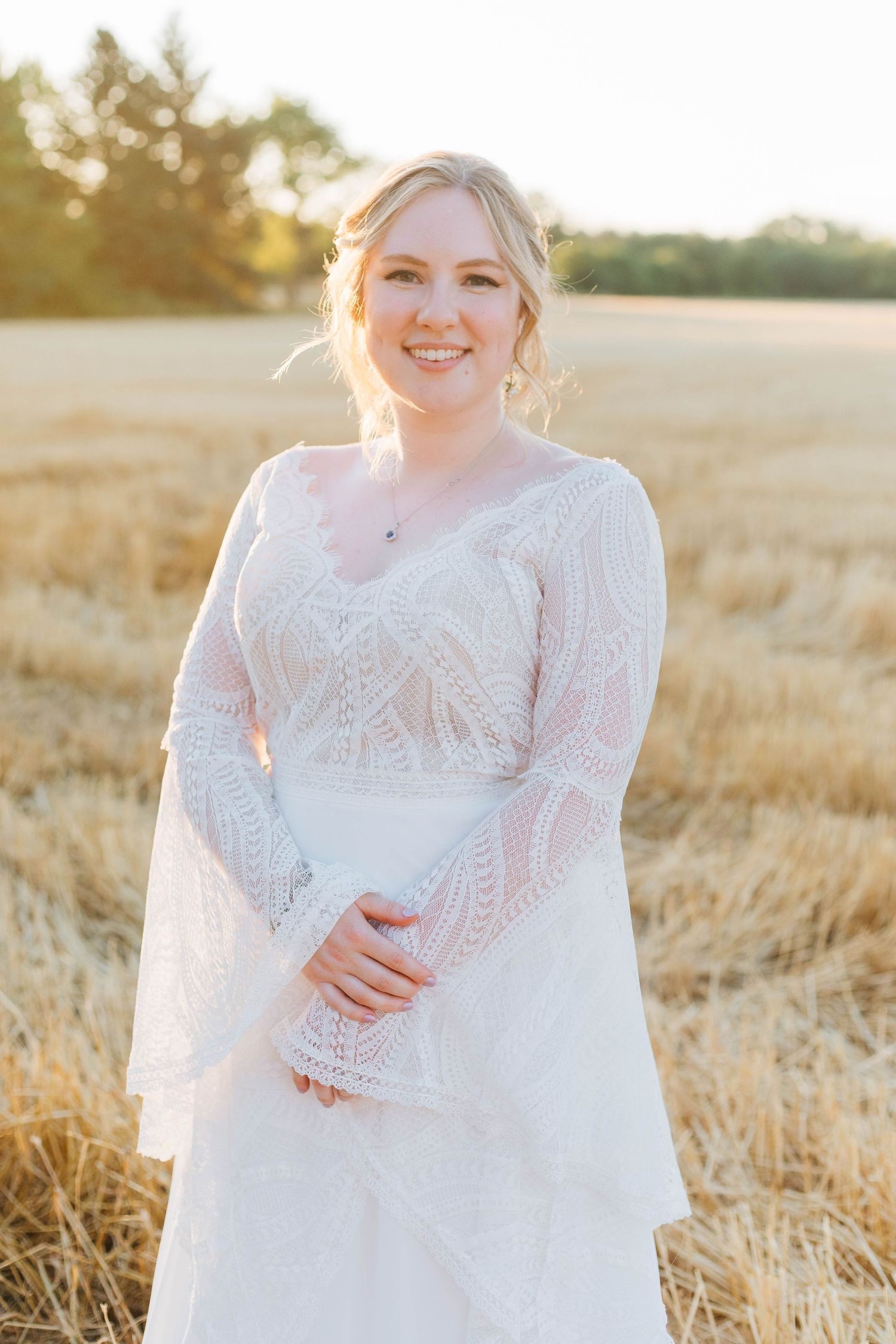 Smiling blonde bride in white lace long-sleeve boho wedding dress standing in golden wheat field at sunset.