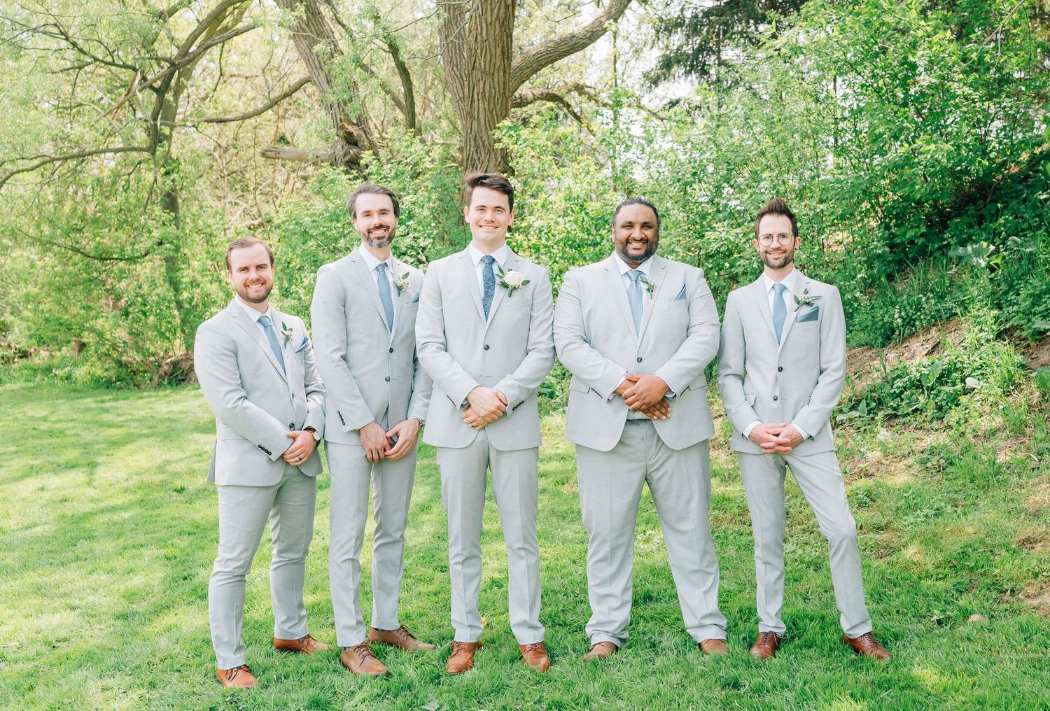 Five groomsmen in matching light gray suits and blue ties pose together on green lawn under large tree.
