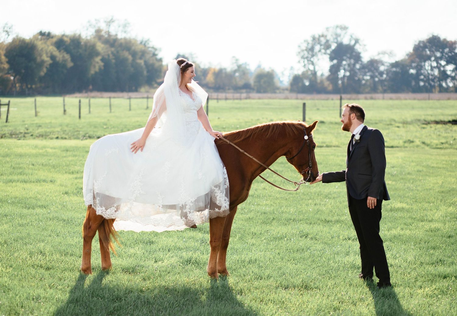 Bride in white gown sits on chestnut horse while groom in black suit stands beside her on green farm field.
