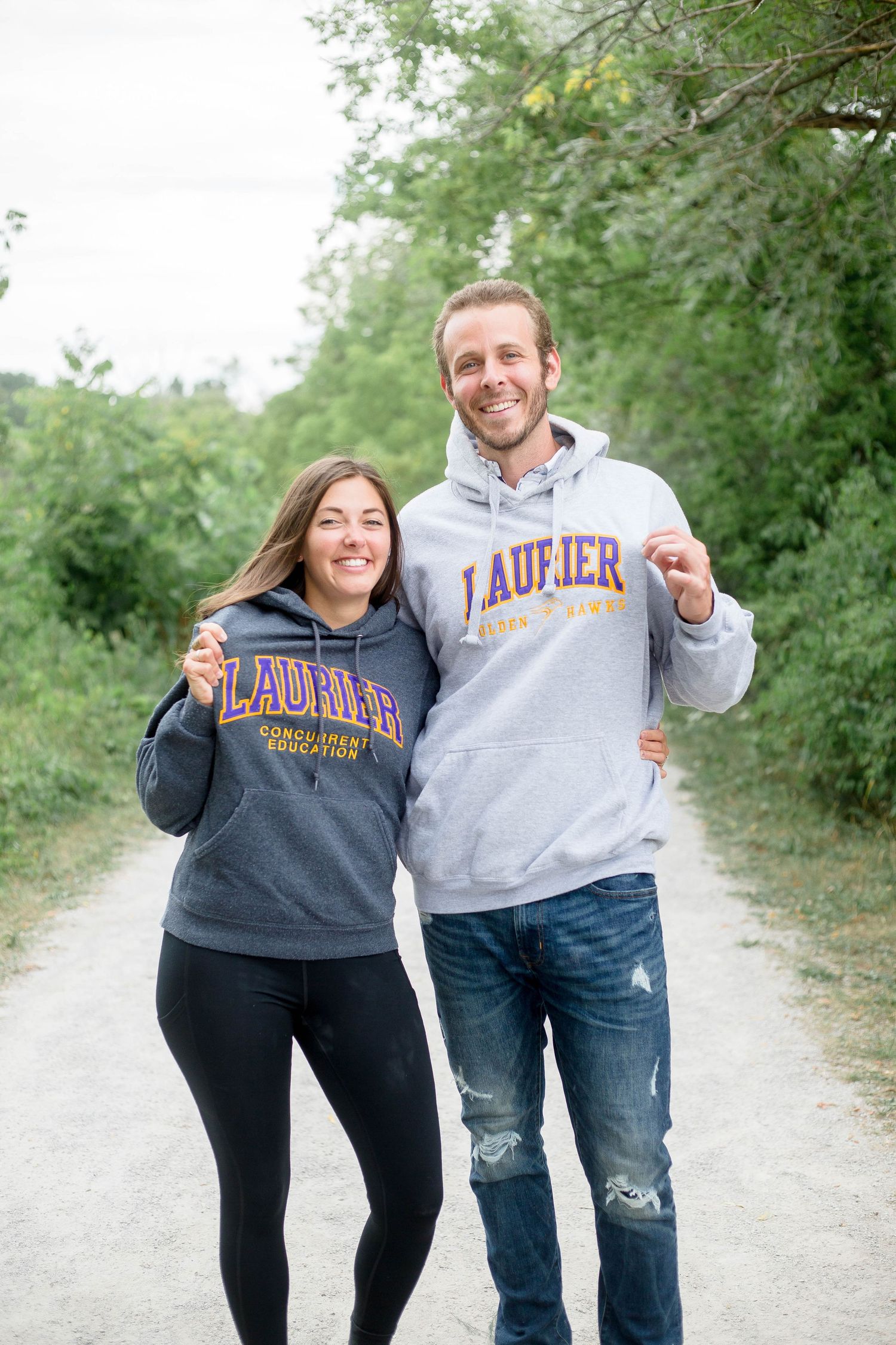 Couple laughing and walking together wearing matching Laurier University hoodies on a green nature trail.
