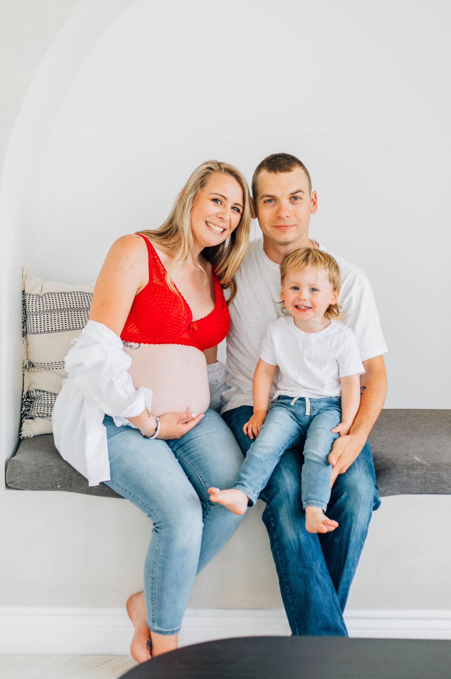 Happy family with pregnant mother, father, and toddler son sitting together on bench smiling.