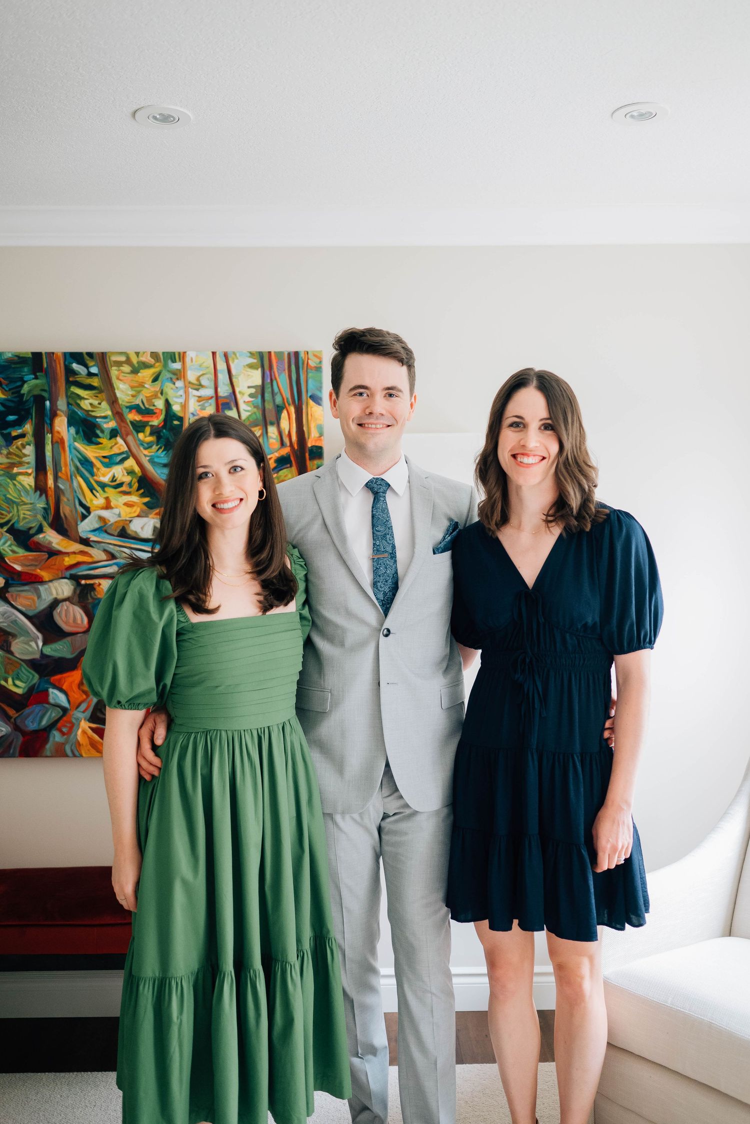 Groom in light gray suit and blue tie posing with two smiling women in elegant dresses.