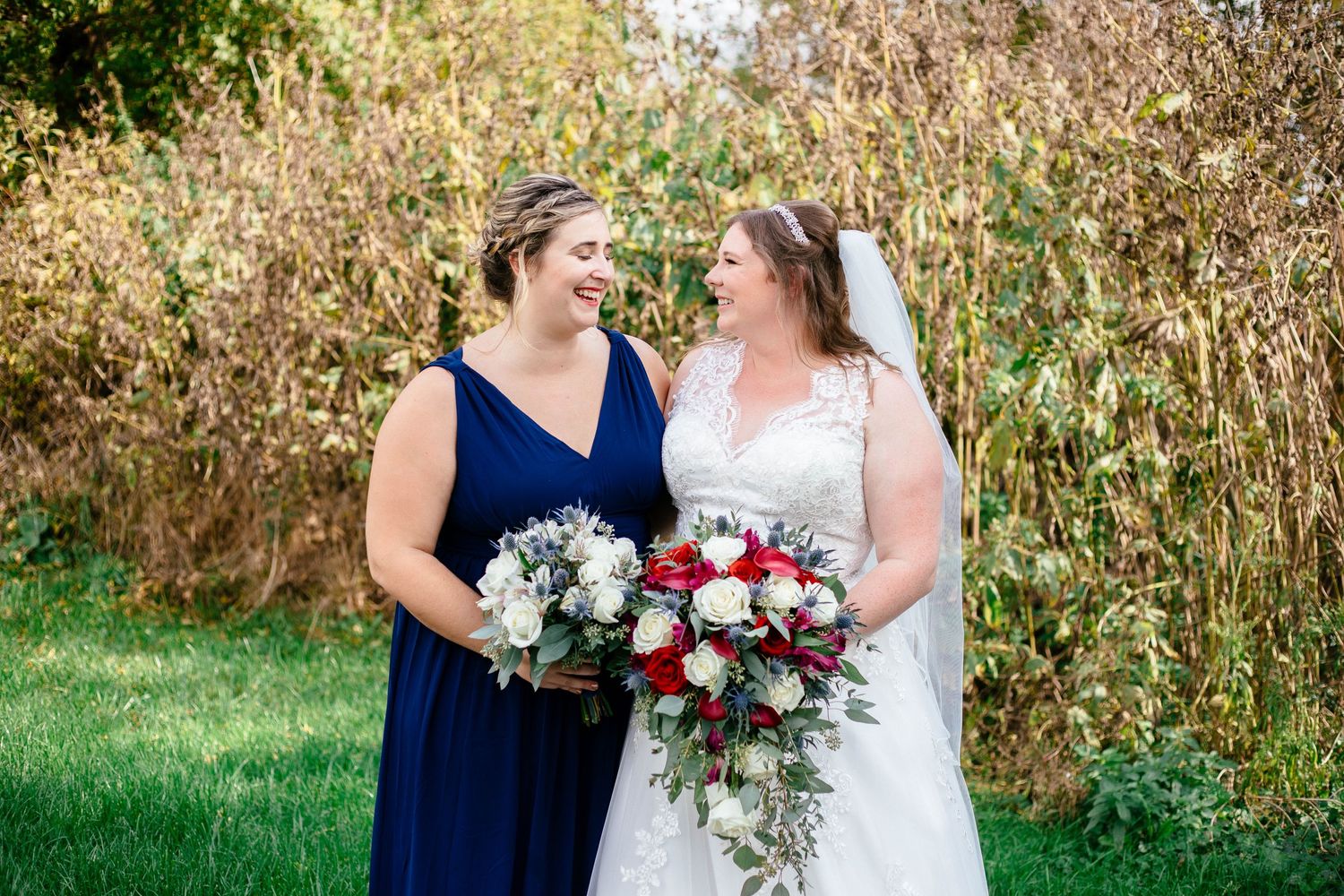 Bride and bridesmaid in navy dress smile warmly at each other while holding wedding bouquets outdoors.