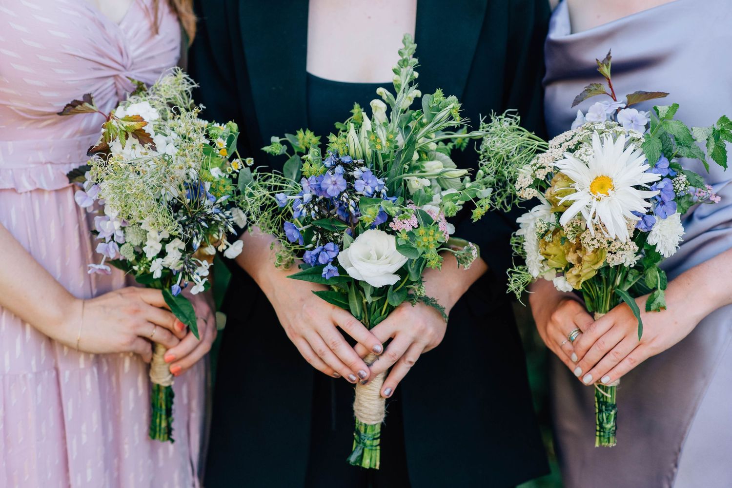 Three women display wildflower bouquets of white daisies, blue flowers, and greenery at a wedding.