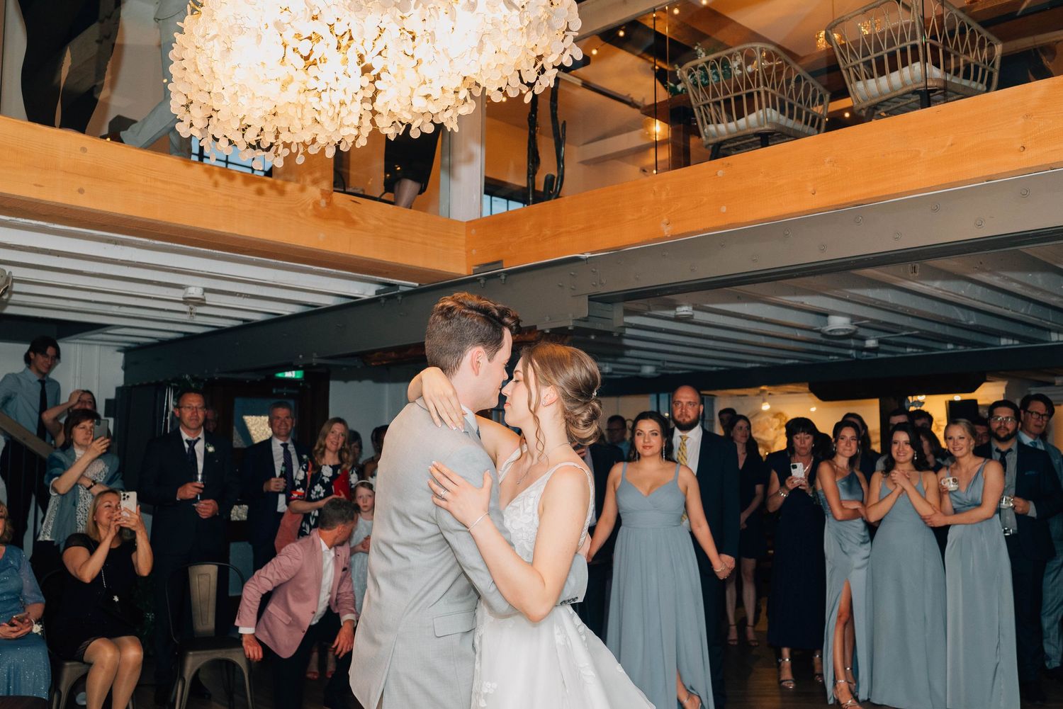 Bride and groom sharing first kiss on dance floor surrounded by bridesmaids in sage dresses.