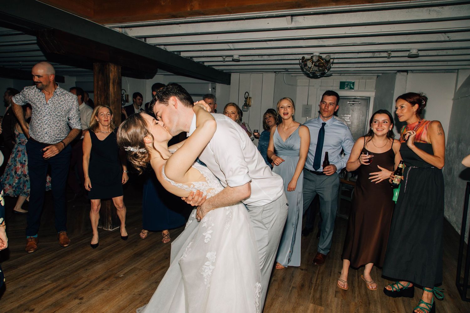 Bride and groom share a passionate dip kiss on the dance floor surrounded by guests.