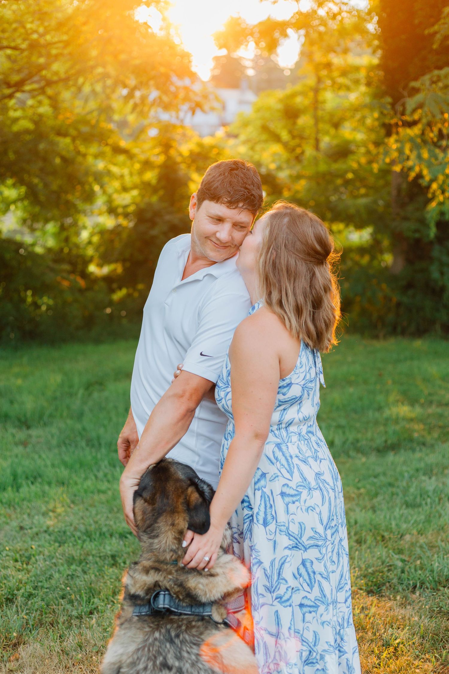 Couple with German Shepherd dog in golden sunset light, woman in blue floral dress kissing mans cheek outdoors.